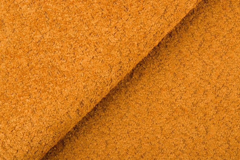 Op de afbeelding: Close-up van gevouwen, getextureerde stof in een warme, goudoranje tint. Het materiaal heeft een dichte, noppenachtige oppervlakte, wat wijst op een pluche of getextureerde textiel. De plooien van de stof cre&euml;ren schaduwen en diepte.