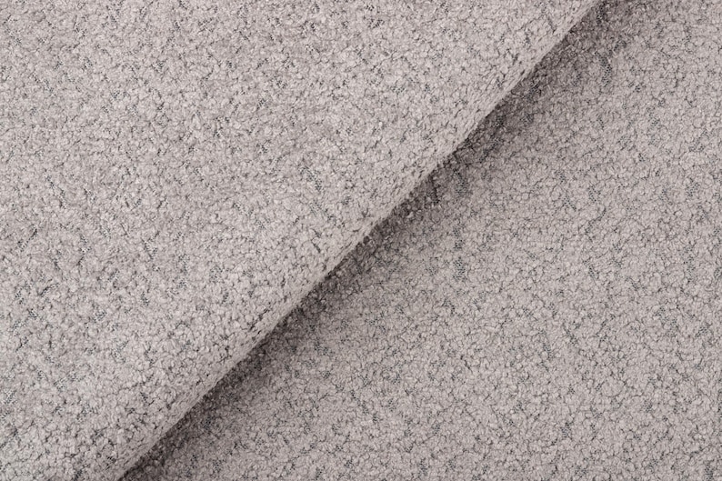 Op de afbeelding: Close-up van een lichtgrijze stof met een getextureerd, noppig oppervlak. De stof is gevouwen en onthult een diagonale vouw. Het materiaal lijkt zacht en kan worden gebruikt voor bekleding of kleding.
