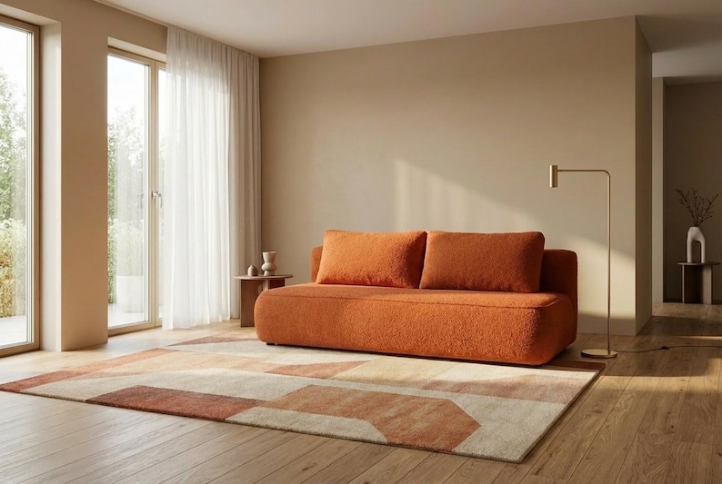 Op de afbeelding: Een oranje bank met twee bijpassende kussens staat op een geometrisch patroon tapijt in een woonkamer. Het tapijt heeft beige, bruine en oranje tinten. Een gouden vloerlamp en een kleine bijzettafel zijn ook zichtbaar.