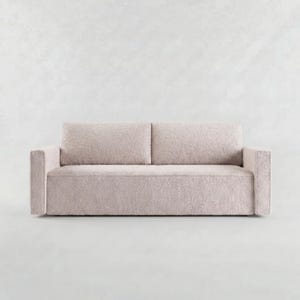 Op de afbeelding: Een lichtbeige bank met een gestructureerde bekleding in bouclé-stijl. Het meubel heeft een rechthoekig silhouet met twee rugkussens, één lang zitkussen en twee brede, platte armleuningen aan weerszijden van het frame.