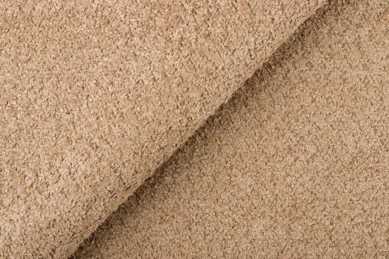 Op de afbeelding: Close-up van een getextureerde stof in een warme, neutrale beige kleur. Het materiaal heeft een noppige, licht onregelmatige oppervlakte, met een gevouwen gedeelte dat een diagonale lijn cre&euml;ert. De stof ziet er zacht en uitnodigend uit.