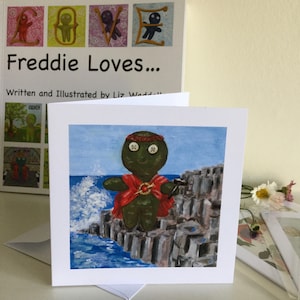 Può includere: Un biglietto d'auguri con un omino di pan di zenzero verde con una veste rossa e una corona in piedi su una scogliera che si affaccia sul mare. Il biglietto ha il testo "Freddie Loves..." sul davanti.