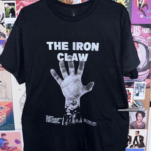 Op de afbeelding: Zwart T-shirt met een witte afbeelding van een hand met de tekst "THE IRON CLAW" erboven. De afbeelding bevat ook de namen "Zac Efron", "Jeremy Allen White", "Harris Dickinson", "Stanley Tucci" en "Holt McCallany".