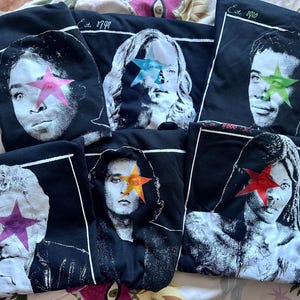 May include: Six black t-shirts featuring high-contrast, monochromatic portraits of individuals. Each portrait is overlaid with a vibrant, solid-coloured star covering one eye. The shirts include white rectangular borders and text such as Est. 1799, Est. 1910, and Est. 4000 BCE.