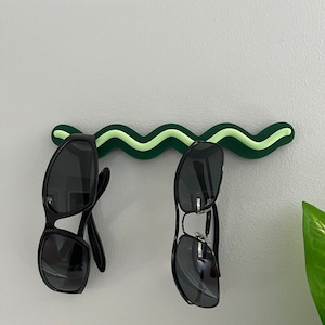 May include: A green and light green wavy sunglasses holder mounted on a light gray wall. Two pairs of black sunglasses are hanging from the holder. A green plant is visible in the background.