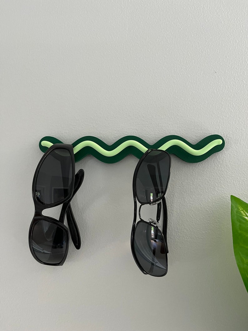 May include: A green and light green wavy sunglasses holder mounted on a light gray wall. Two pairs of black sunglasses are hanging from the holder. A green plant is visible in the background.