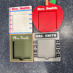 May include: Four teacher-themed desk organizers. One is apple-shaped, red with a green leaf and stem. Another is a white frame with blue and pink stripes. The third is olive green, and the fourth is gray with calculator symbols. Each has "Mrs. Smith" printed on it.