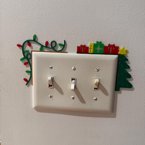 May include: A decorative Christmas light switch cover. The cover is white with three light switches. The top of the cover is decorated with a green garland with red lights, green and red gift boxes, a green Christmas tree, and a yellow star.