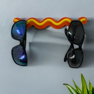 May include: A novelty sunglasses holder shaped like a hot dog, with a wavy yellow and orange design. Two pairs of sunglasses hang from the holder, one with blue reflective lenses and the other with dark lenses. The holder is mounted on a light blue wall.
