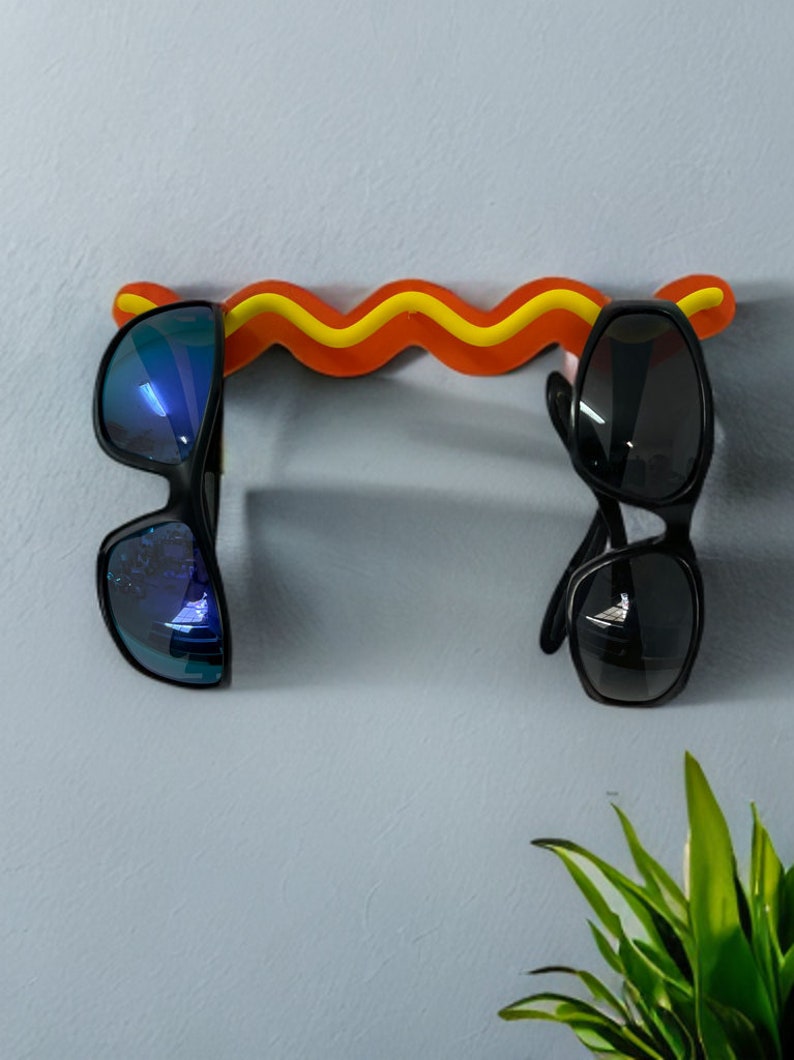 May include: A novelty sunglasses holder shaped like a hot dog, with a wavy yellow and orange design. Two pairs of sunglasses hang from the holder, one with blue reflective lenses and the other with dark lenses. The holder is mounted on a light blue wall.