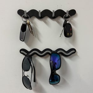 May include: Two black wavy key and sunglass holders mounted on a white wall. The top holder has keys and a car remote hanging from it. The bottom holder has two pairs of sunglasses hanging from it.