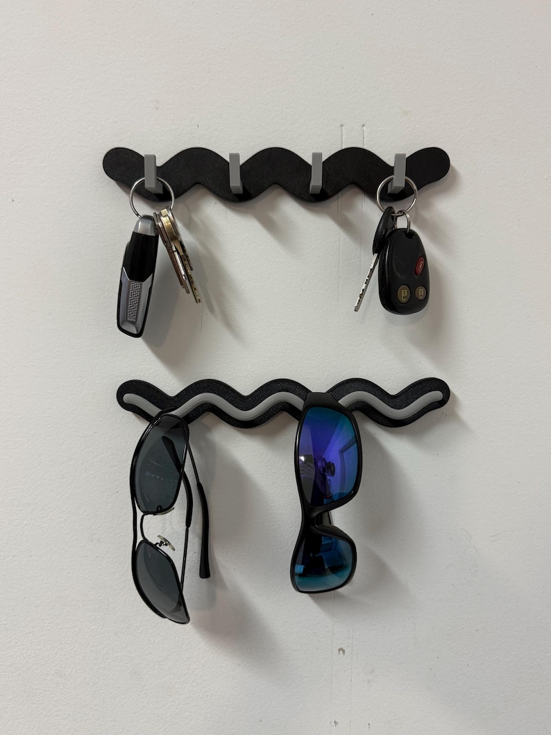 May include: Two black wavy key and sunglass holders mounted on a white wall. The top holder has keys and a car remote hanging from it. The bottom holder has two pairs of sunglasses hanging from it.