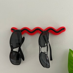 May include: Two pairs of black sunglasses hang from a red and black wavy wall-mounted sunglasses holder. The sunglasses have dark lenses and black frames. The holder is mounted on a white wall.