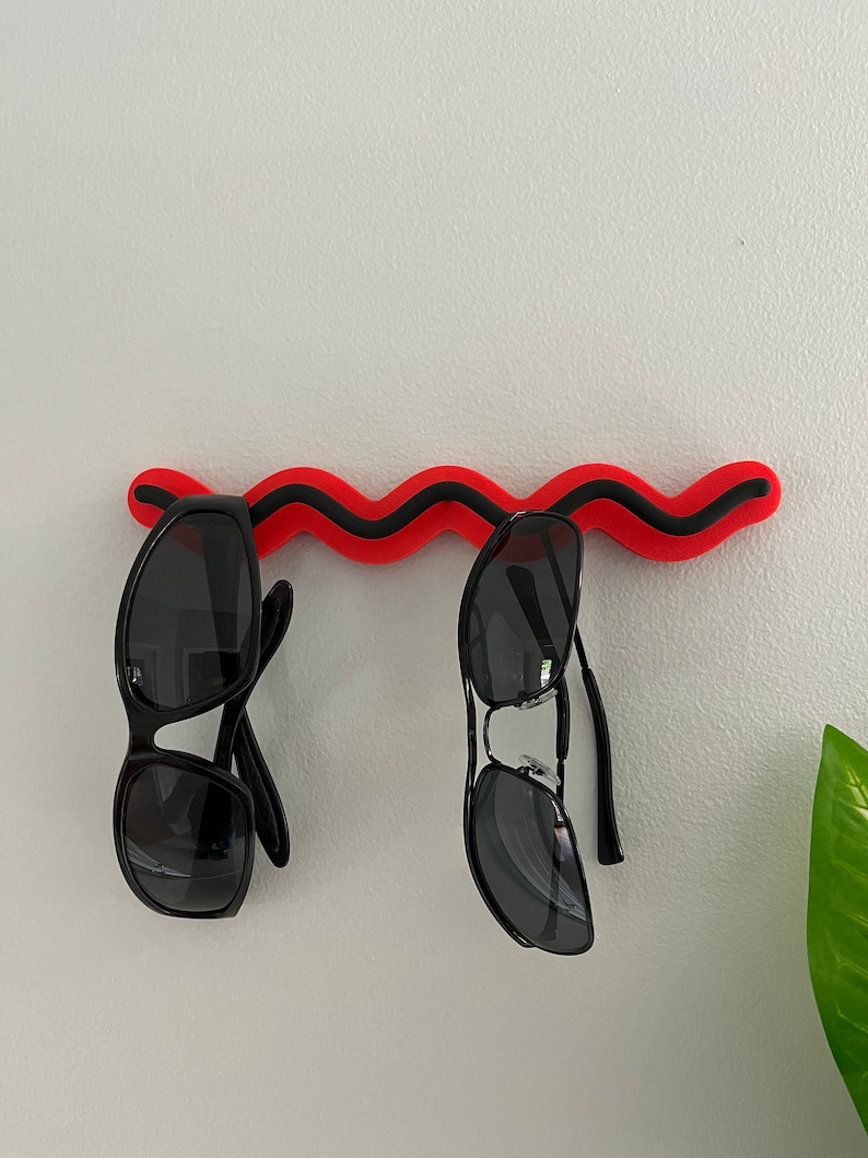 May include: Two pairs of black sunglasses hang from a red and black wavy wall-mounted sunglasses holder. The sunglasses have dark lenses and black frames. The holder is mounted on a white wall.