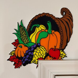 May include: A colorful cornucopia decoration overflowing with autumn harvest items. The brown horn-shaped basket is filled with a yellow squash, orange pumpkin, red apple, purple grapes, and yellow corn. Red, yellow, and green leaves complete the fall theme.
