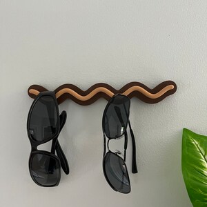 May include: A brown and tan wavy sunglasses holder mounted on a white wall, with two pairs of black sunglasses hanging from it. The holder has a unique, decorative design. A green plant is visible in the background.