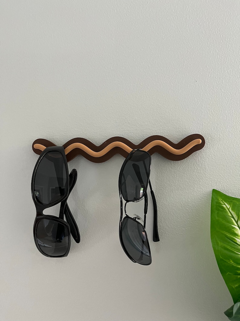 May include: A brown and tan wavy sunglasses holder mounted on a white wall, with two pairs of black sunglasses hanging from it. The holder has a unique, decorative design. A green plant is visible in the background.