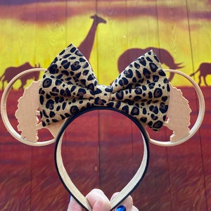 May include: A pair of brown and tan mouse ears with a leopard print bow. The ears are shaped like trees and the bow is tied in the center. The background is a brown wooden wall with a sunset painted on it.