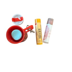 May include: A red keychain with a small red basket, a blue ball, and a white lid. Next to it are two lip balm tubes, one yellow and one with a pinkish-brown tint. The lip balm tubes are labeled with text.