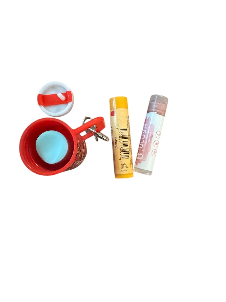 May include: A red keychain with a small red basket, a blue ball, and a white lid. Next to it are two lip balm tubes, one yellow and one with a pinkish-brown tint. The lip balm tubes are labeled with text.