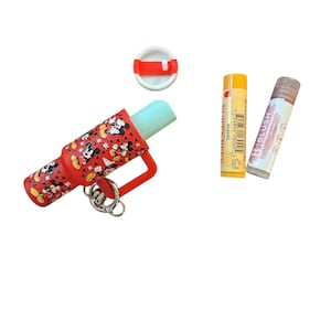 May include: A red lip balm holder with a Mickey Mouse pattern, a white and red lid, and two lip balms. One lip balm is yellow with a barcode, and the other is pink with the word "BLUSH".