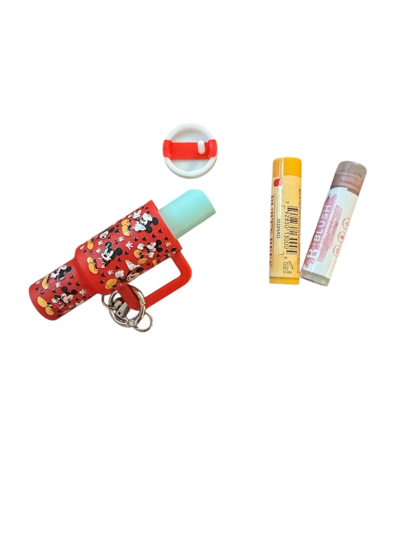 May include: A red lip balm holder with a Mickey Mouse pattern, a white and red lid, and two lip balms. One lip balm is yellow with a barcode, and the other is pink with the word "BLUSH".