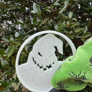 Pode incluir: Uma faixa de cabe&ccedil;a de pl&aacute;stico branco com um recorte do personagem Jack Skellington do filme O Estranho Mundo de Jack. A faixa de cabe&ccedil;a est&aacute; sendo segurada por um tecido verde com um padr&atilde;o de teia de aranha.