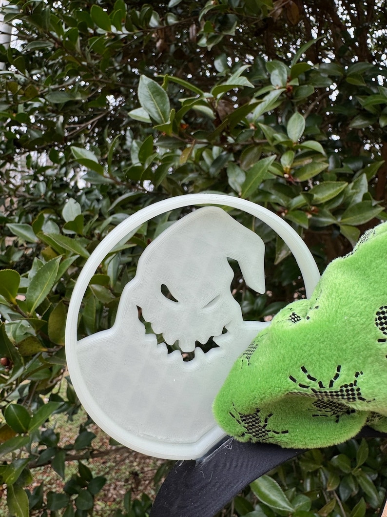 Pode incluir: Uma faixa de cabe&ccedil;a de pl&aacute;stico branco com um recorte do personagem Jack Skellington do filme O Estranho Mundo de Jack. A faixa de cabe&ccedil;a est&aacute; sendo segurada por um tecido verde com um padr&atilde;o de teia de aranha.