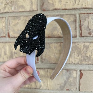 May include: A white headband with a black, crystal-studded mask-shaped embellishment. The mask has eye and mouth cutouts. The headband is held against a brick wall.