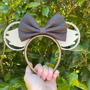 May include: White mouse ears with a black bow, featuring a silhouette of a hand on each ear.