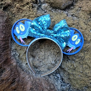 May include: A headband featuring two circular blue ears, each displaying a cartoon snake face with yellow eyes and a pink tongue. A large, bright blue sequined bow is centered between the ears. The headband frame is a simple, solid blue band.