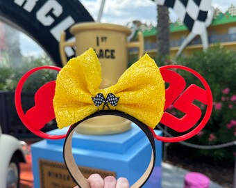 May include: A black headband with red Mickey Mouse ears featuring the number 25. The headband has a yellow sequined bow with two checkered flags. The background includes a trophy and a race car.
