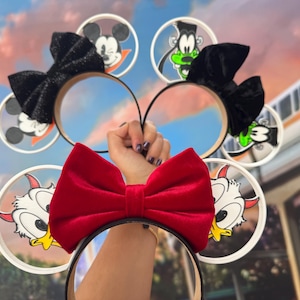 May include: A collection of character-themed ear headbands. The headbands feature black bows and character designs, including Mickey Mouse, Goofy, and Donald Duck. The red bow headband is in the foreground.