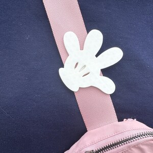 Puede incluir: Un bolso de hombro rosa y blanco con un dije de guante de Mickey Mouse blanco sujeto a la correa.