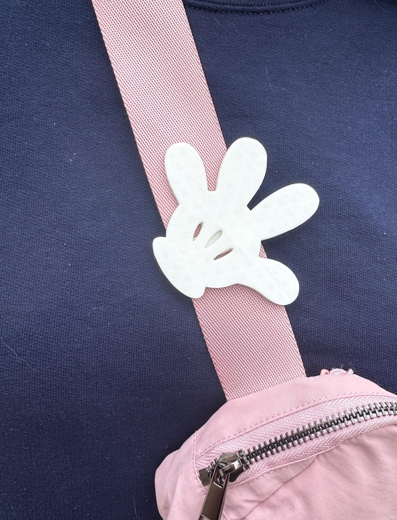Puede incluir: Un bolso de hombro rosa y blanco con un dije de guante de Mickey Mouse blanco sujeto a la correa.