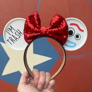 May include: Red sequin bow headband with white ears. One ear has the text "I'M TRASH" and the other ear has a cartoon face with blue eyes, a red mouth, and a red line above the eyes.