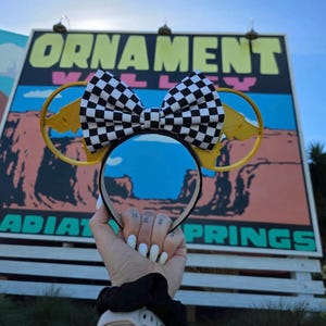 May include: Black headband with yellow Mickey Mouse ears and a checkered bow. The ears have a yellow lightning bolt design. The headband is held in front of a sign that says "Ornament Valley" and "Radiator Springs".