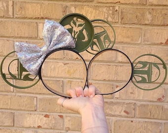 May include: A pair of black headband mouse ears with a silver glitter bow. The ears are decorated with green silhouettes of Star Wars characters, including Boba Fett, Mandalorian, and a Stormtrooper.