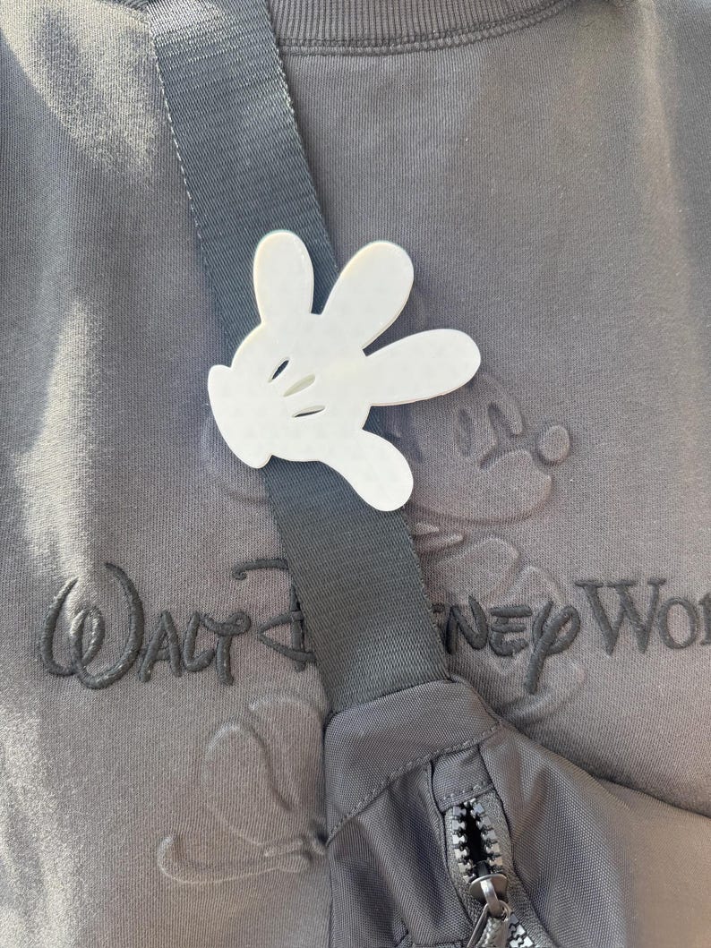 Puede incluir: Una ri&ntilde;onera negra y gris con cierre de cremallera. La ri&ntilde;onera se lleva sobre una sudadera gris con el texto "Walt Disney World" bordado en la parte delantera. Una mano de Mickey Mouse de pl&aacute;stico blanco est&aacute; unida a la correa de la ri&ntilde;onera.