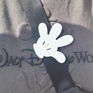 Puede incluir: Un recorte de mano blanca de Mickey Mouse sujeto a una correa negra en una camisa gris con el texto "Walt Disney World" bordado en ella.