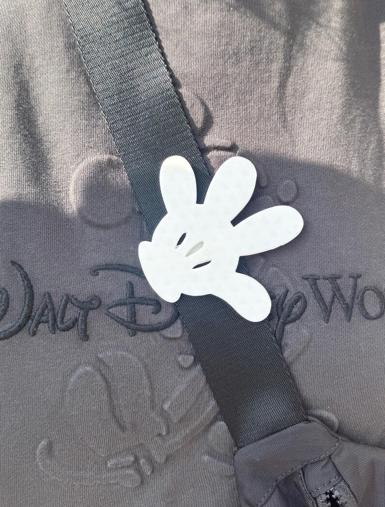 Puede incluir: Un recorte de mano blanca de Mickey Mouse sujeto a una correa negra en una camisa gris con el texto "Walt Disney World" bordado en ella.
