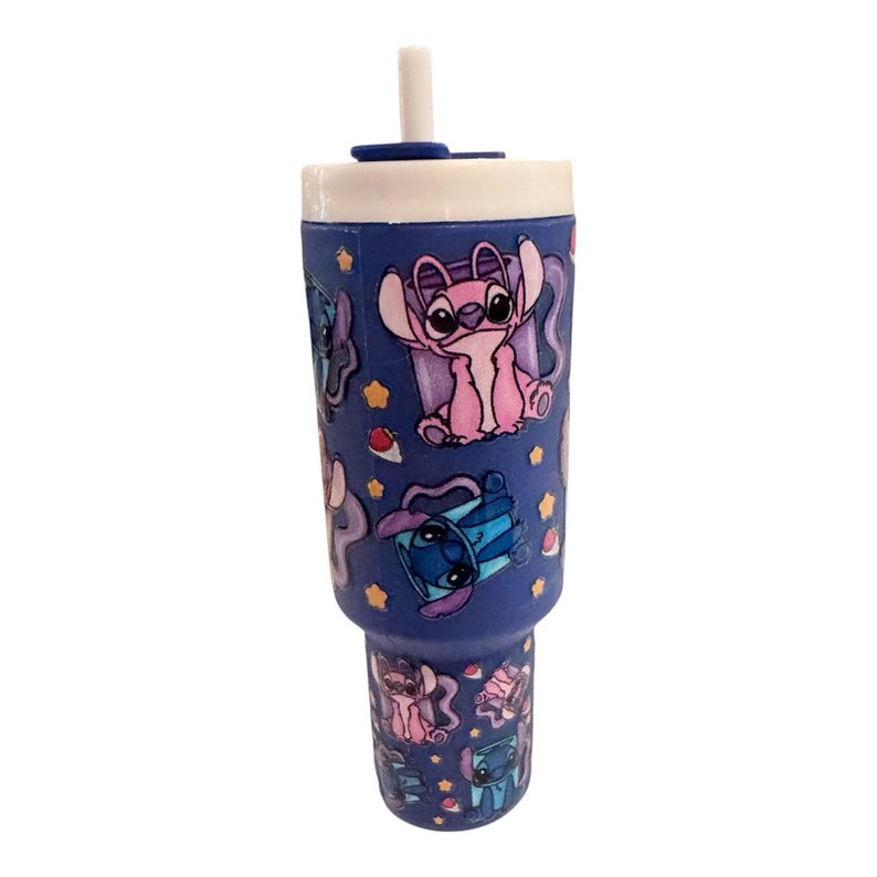 May include: A blue tumbler with a white lid and straw. The tumbler is decorated with cartoon characters in various poses, including a pink and blue alien. The design includes stars and other small shapes.