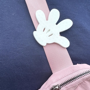 Puede incluir: Una bolsa rosa y azul marino con un dije de mano de Mickey Mouse blanco sujeto a la correa. La bolsa tiene un cierre de cremallera.
