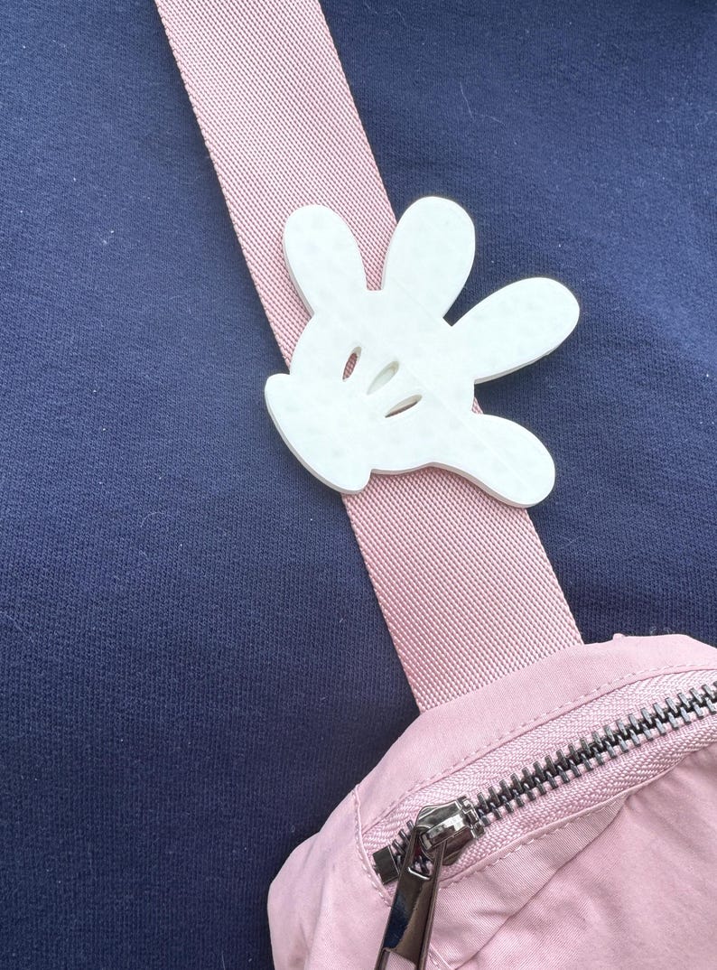 Puede incluir: Una bolsa rosa y azul marino con un dije de mano de Mickey Mouse blanco sujeto a la correa. La bolsa tiene un cierre de cremallera.