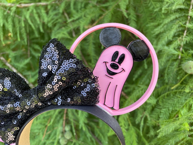 May include: Pink Mickey Mouse ghost headband with black sequin bow. The headband is made of plastic and features a pink ghost with Mickey Mouse ears and a black outline. The bow is made of black sequins and is attached to the headband.