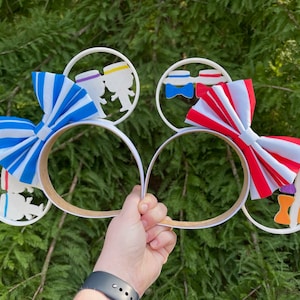 May include: White headband with two large bows, one blue and white striped and the other red and white striped. Each bow is attached to a white circle with a cutout design of two figures holding hands.
