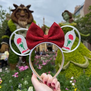 May include: White headband with silver glitter ears featuring a red rose and a glass dome. The headband has a red glitter bow.