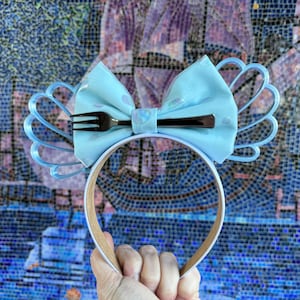 May include: Light blue headband with a fork-shaped bow and a seashell design. The headband is white with a brown band.