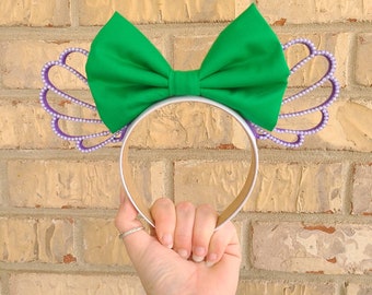 May include: Purple and silver headband with a green bow. The headband features a design of a crown with pearl accents.