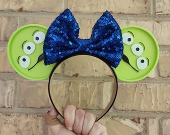 May include: Green and white character ears with three eyes and a mustache, with a blue sparkly bow on top. The ears are attached to a black headband.